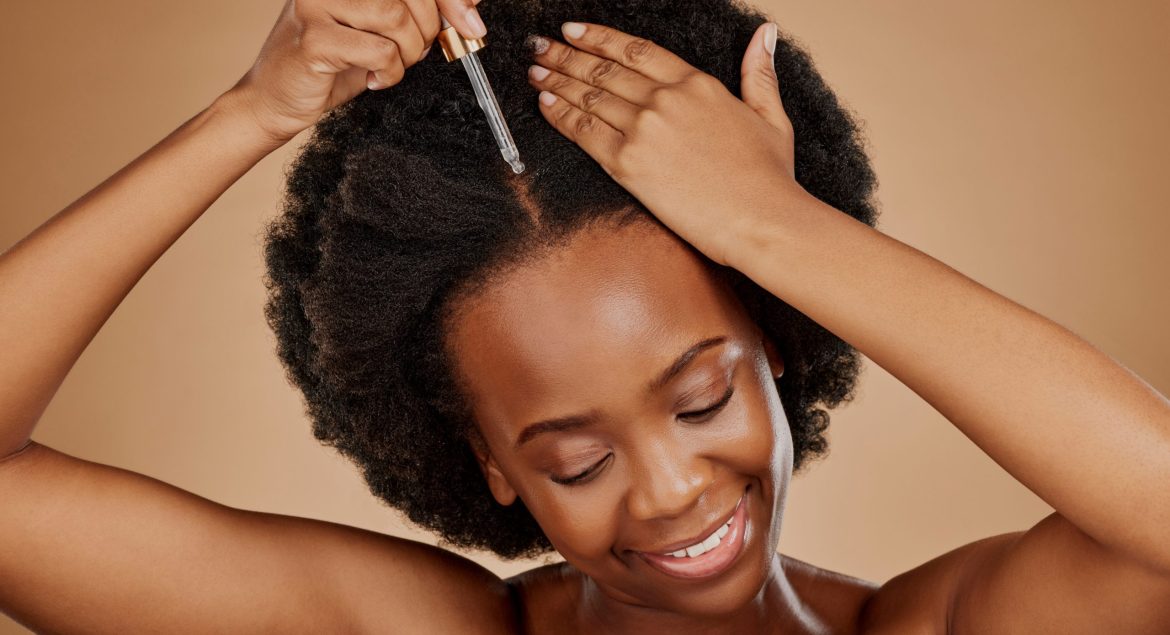 Oil serum, hair care or black woman with afro in studio on brown background for a healthy scalp. Smile, pipette or natural African girl at hairdresser salon for hairstyle treatment or beauty makeover.